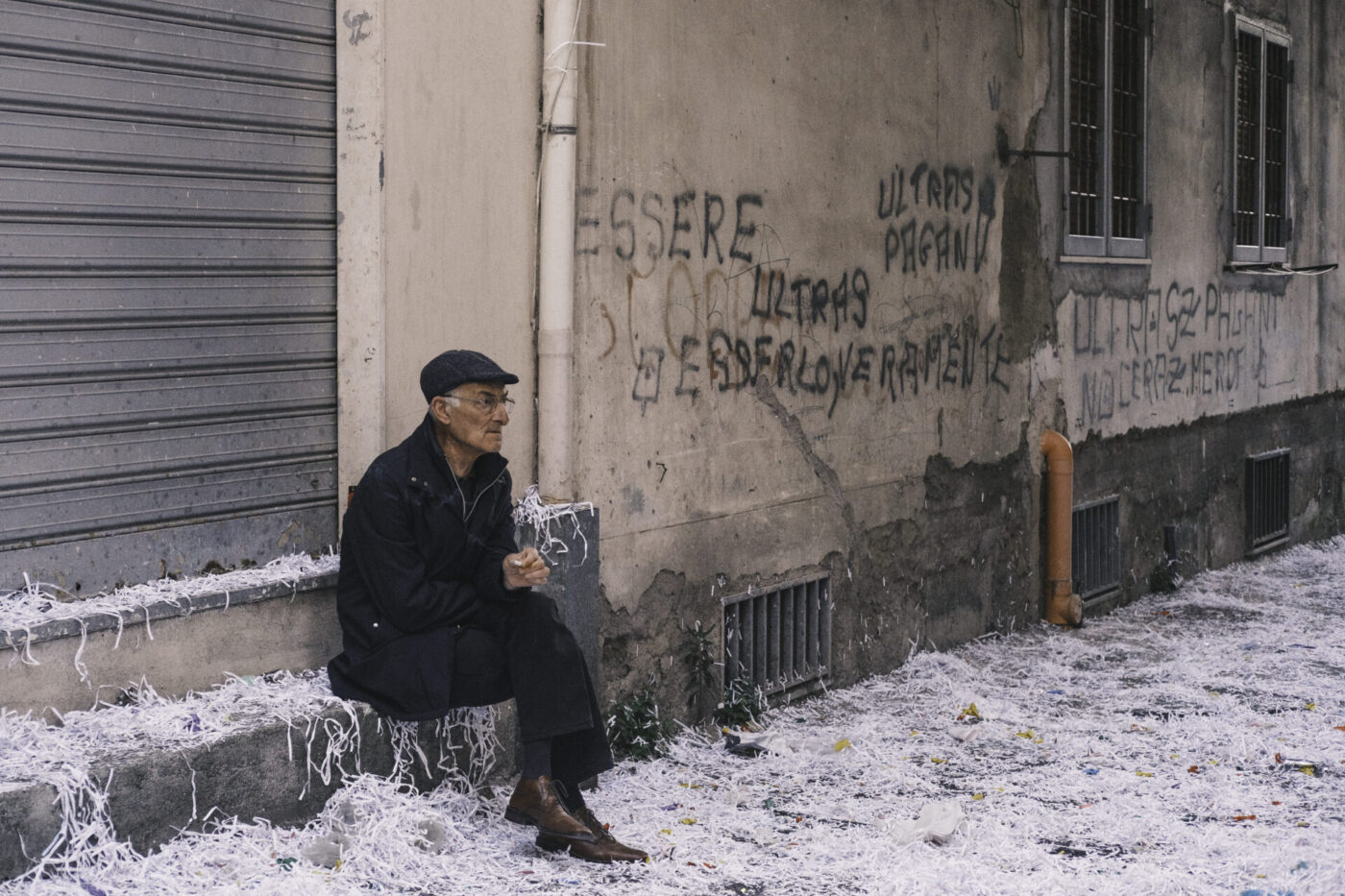 Un uomo anziano con un berretto piatto siede accanto a graffiti, carta stracciata intorno a lui. Ha un'aria pensierosa e tiene dei fogli tra le mani.