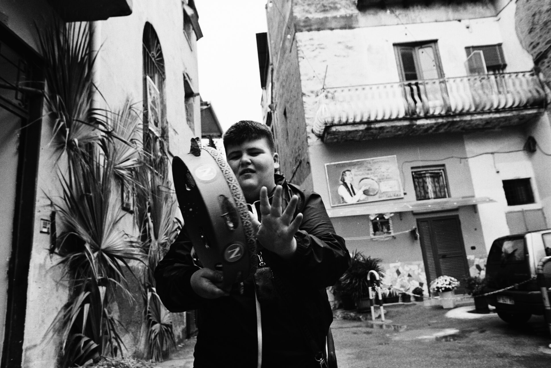 Un ragazzo suona il tamburello in una strada, con la mano a mezz'aria; dietro di lui vecchi edifici, piante, un murale. Foto in bianco e nero.