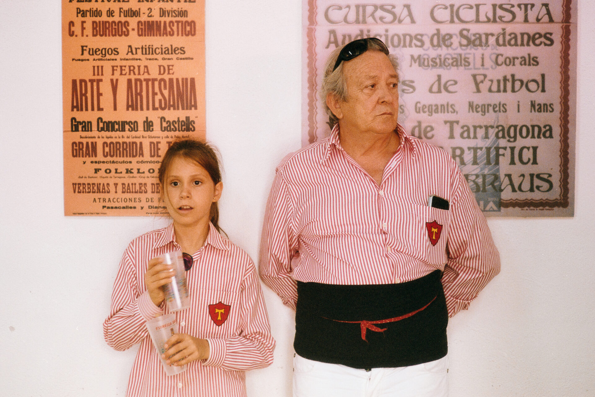Un uomo anziano e una ragazza con camicie a righe bianche e rosse abbinate stanno insieme; la ragazza ha una tazza, alle loro spalle poster d'epoca.