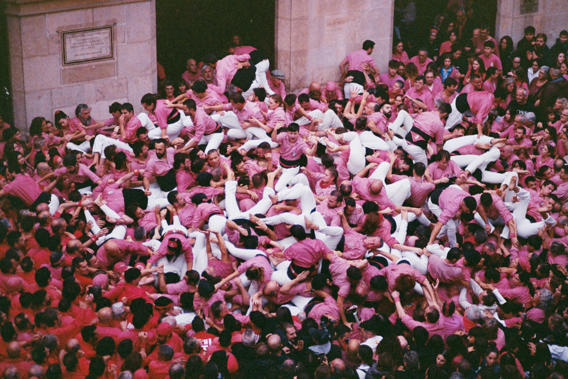 Una folla vivace in camicie rosa e pantaloni bianchi forma un ammasso caotico a un festival, osservata da spettatori in abiti rossi.