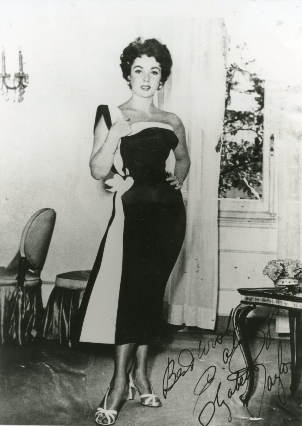 Una donna composta in un abito nero senza spalline con accenti bianchi si trova in una stanza elegante; foto firmata con tendaggi, sedie, tavolo.