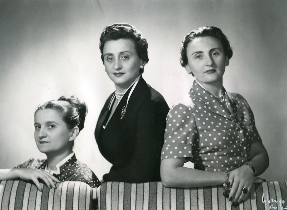 Tre donne in abiti di metà secolo, due a fantasia, siedono e stanno in piedi accanto a un divano a righe, posando con calma per un ritratto in bianco e nero.