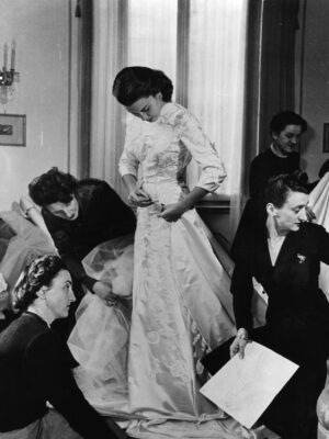 A woman stands in a fitting room as others adjust her ornate dress; someone holds a sketch, tall windows behind them.