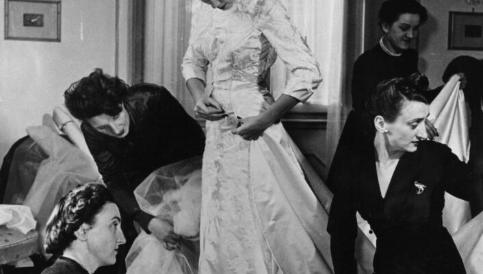 A woman stands in a fitting room as others adjust her ornate dress; someone holds a sketch, tall windows behind them.