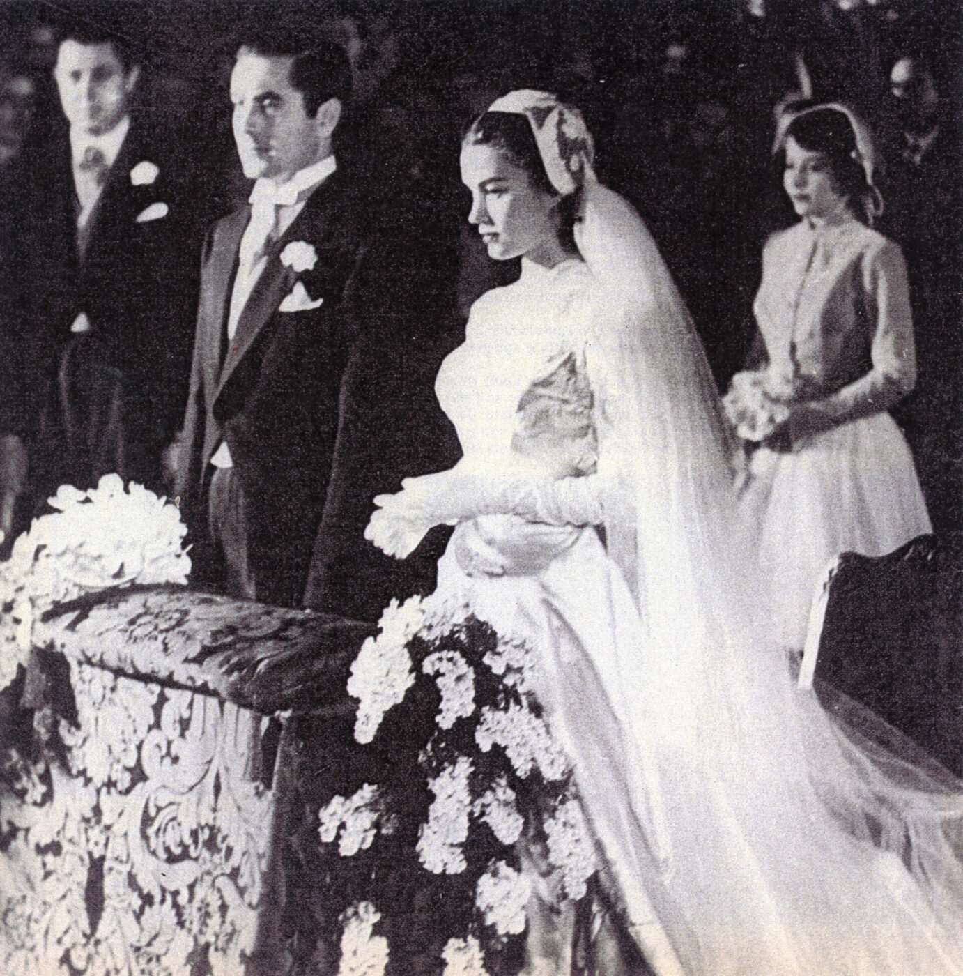 Una sposa in abito bianco e velo sta con lo sposo all'altare; le persone vicine tengono dei fiori in un ambiente elegante e tradizionale.