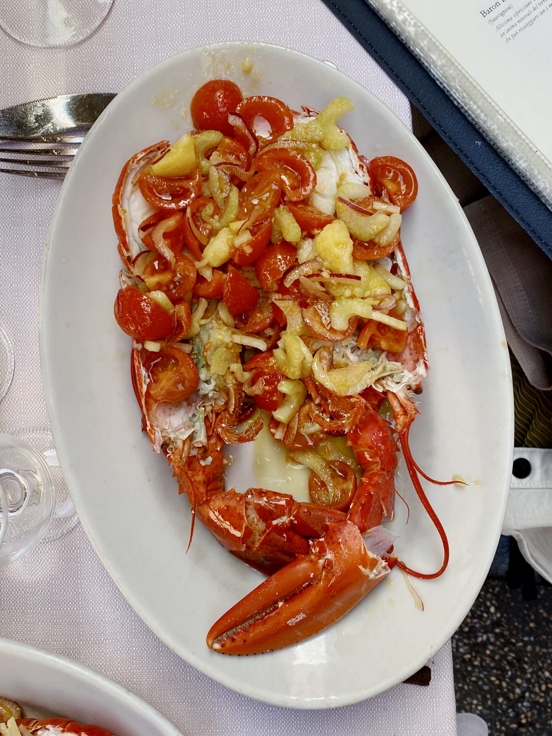 A cooked lobster split in half, topped with cherry tomato salad and onions, sits on a white plate beside utensils and a menu.
