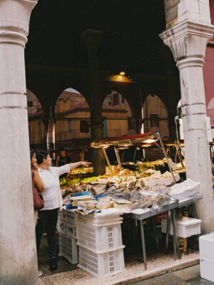 Two women at a busy market stall of seafood and veggies; one points as stone arches and historic buildings frame the scene.