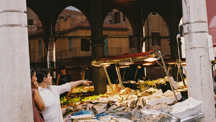 Two women at a busy market stall of seafood and veggies; one points as stone arches and historic buildings frame the scene.