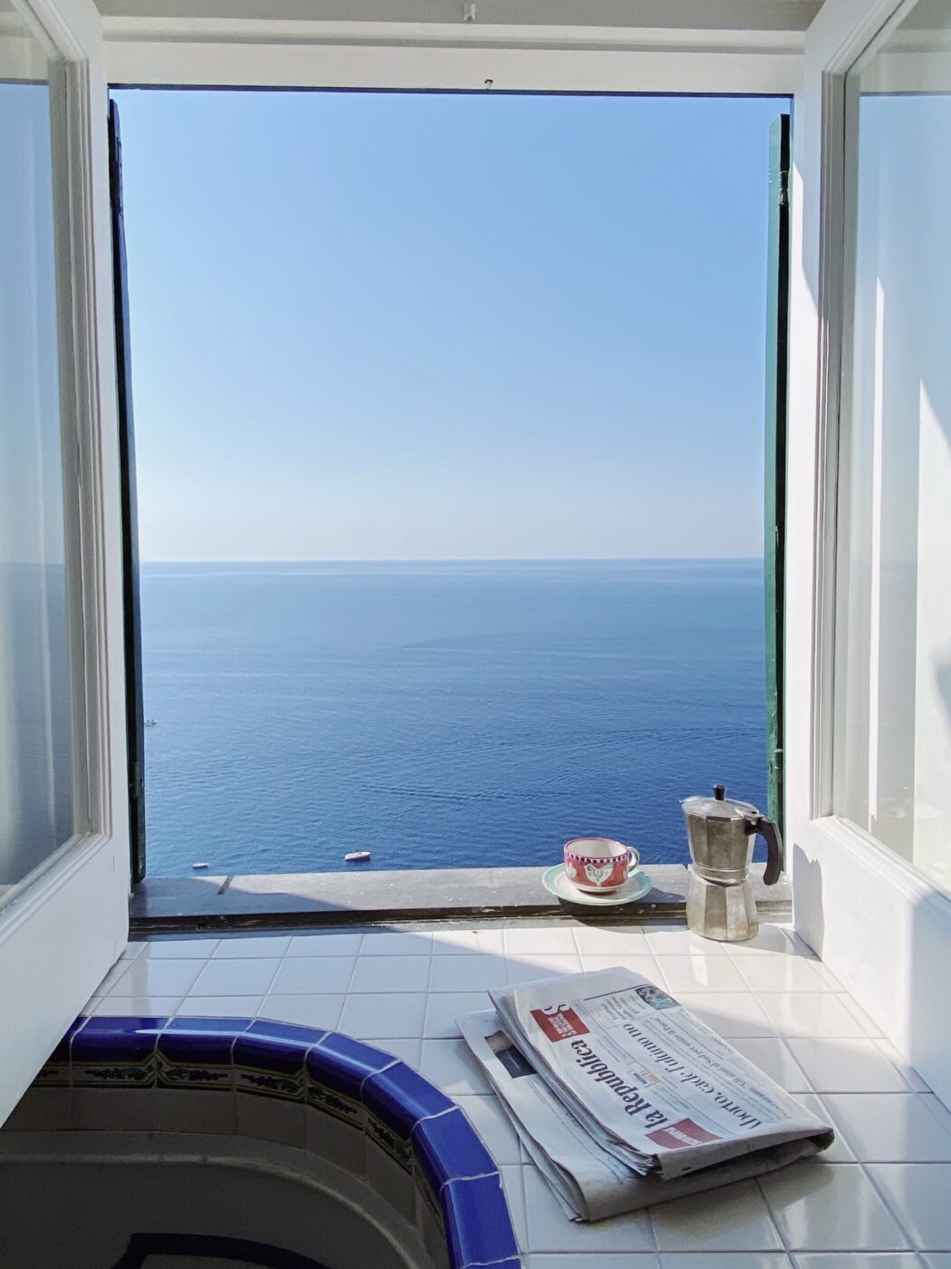 Una finestra illuminata dal sole si apre su un mare azzurro e calmo; sul davanzale, un caffè con un giornale piegato crea un'atmosfera serena.