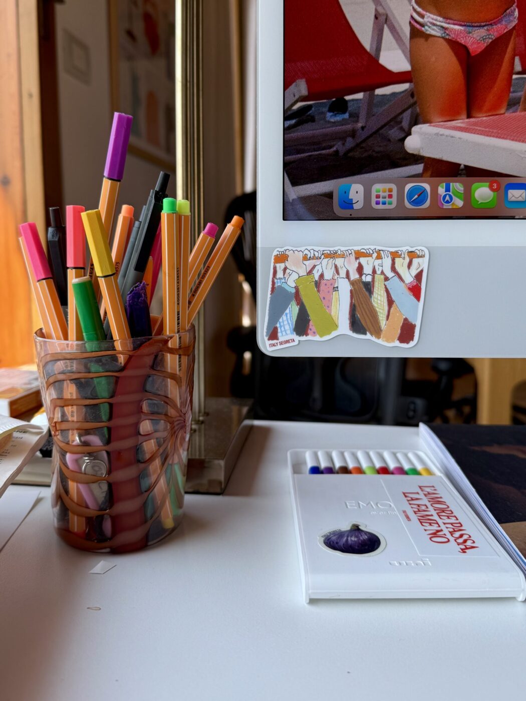A Tavola Sticker Pack - A glass holds colorful pens and markers on a white desk next to an iMac displaying a beach photo. Nearby are colored pencils in a white case and an A Tavola Sticker Pack sticker with hanging clothes is attached to the monitor.