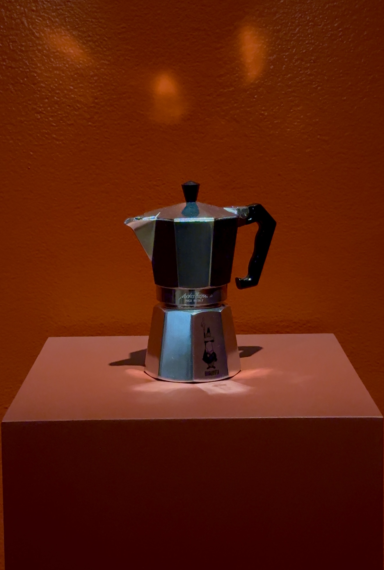 Una macchina da caffè espresso argentata su un piedistallo rosa si trova davanti a una parete arancione, con un'illuminazione drammatica che proietta ombre e riflessi audaci.