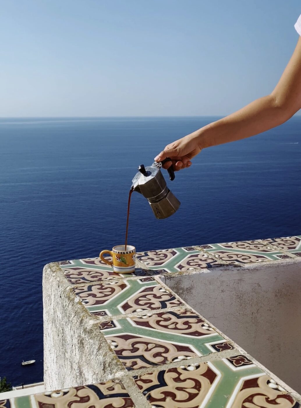 Una persona versa il caffè da una caffettiera moka in una tazza gialla su un davanzale piastrellato, affacciato su un vasto mare blu sotto un cielo limpido.