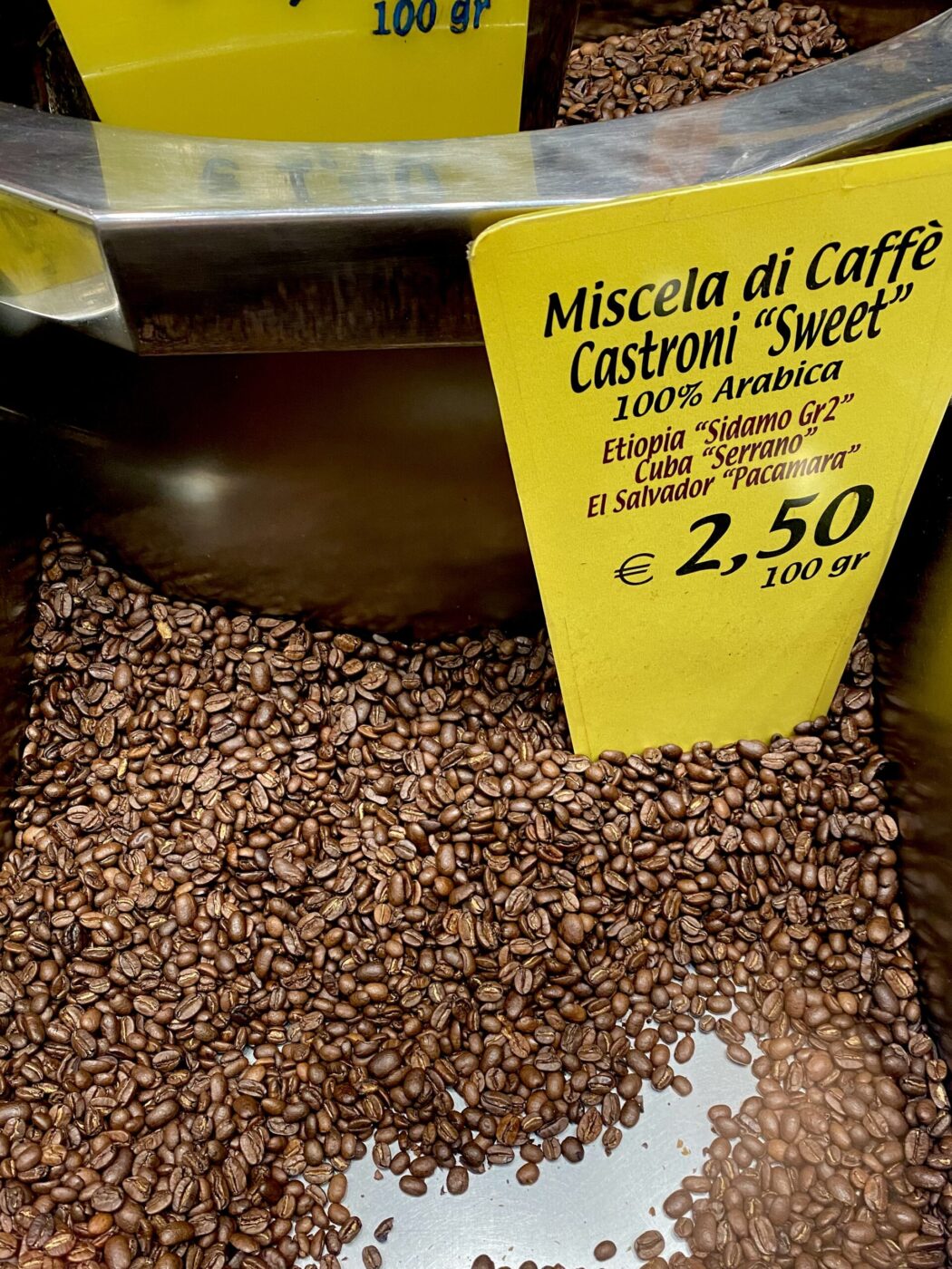Un contenitore metallico di chicchi di caffè tostati con un'etichetta gialla che riporta le informazioni sulla miscela e il prezzo di 2,50 €/100g; include etiopi, cubani e salvadoregni.
