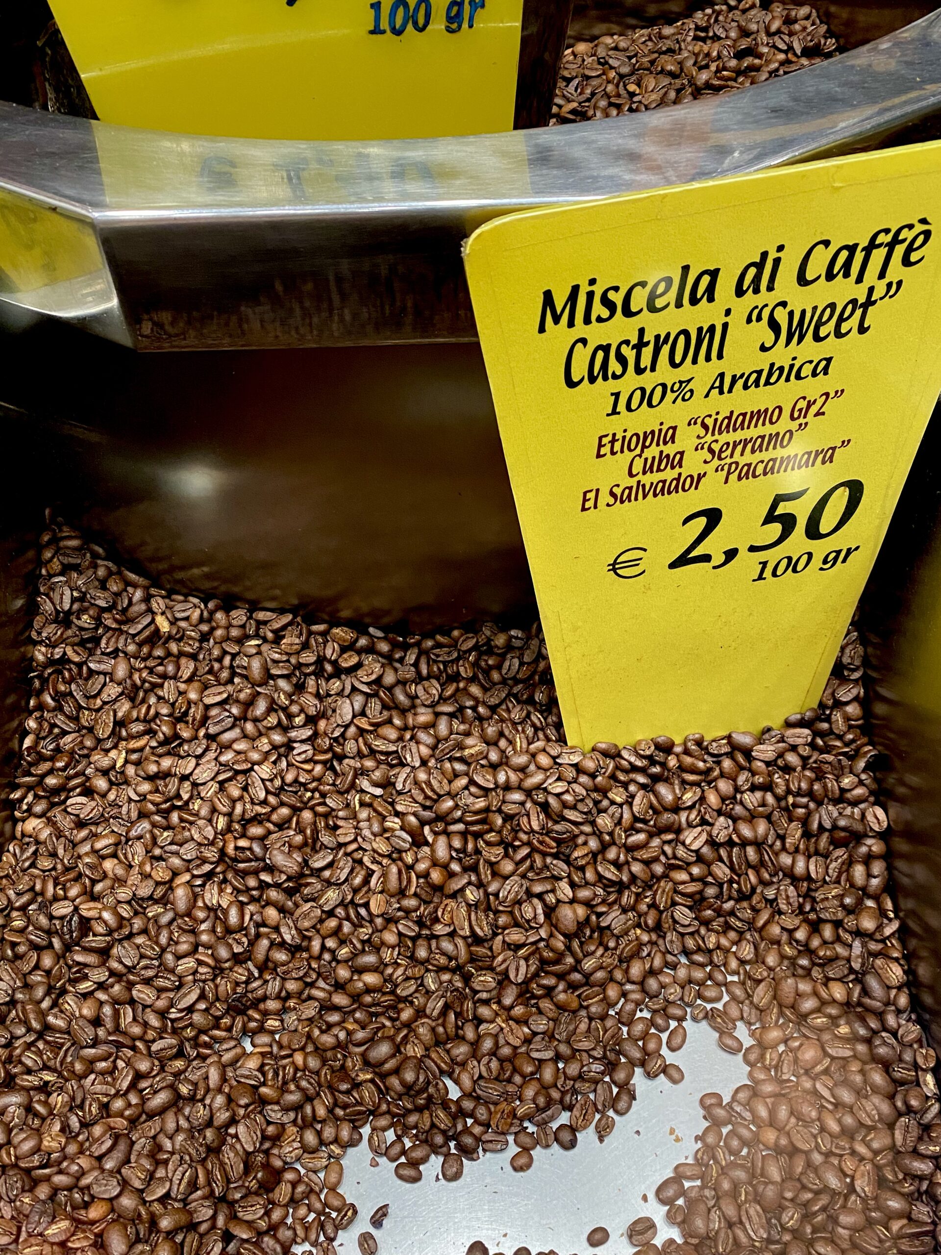 Un contenitore metallico di chicchi di caffè tostati con un'etichetta gialla che riporta le informazioni sulla miscela e il prezzo di 2,50 €/100g; include etiopi, cubani e salvadoregni.