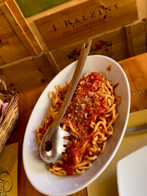 A white oval plate filled with pasta topped with tomato sauce and grated cheese, served with a metal pasta fork, sits on a table with wooden box decor in the background. Just replace [Image Title] with the actual title of your image. For example, if the image title is Spaghetti Dinner, it would be: Spaghetti Dinner - A white oval plate filled with pasta topped with tomato sauce and grated cheese, served with a metal pasta fork, sits on a table with wooden box decor in the background.