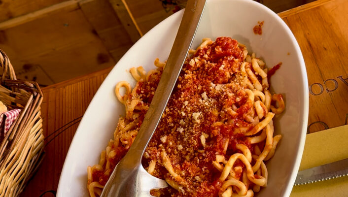 A white oval plate filled with pasta topped with tomato sauce and grated cheese, served with a metal pasta fork, sits on a table with wooden box decor in the background. Just replace [Image Title] with the actual title of your image. For example, if the image title is Spaghetti Dinner, it would be: Spaghetti Dinner - A white oval plate filled with pasta topped with tomato sauce and grated cheese, served with a metal pasta fork, sits on a table with wooden box decor in the background.