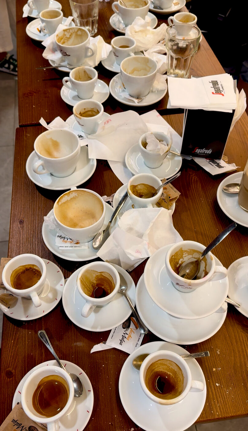 Decine di tazze da caffè vuote e mezze piene, cucchiai usati, tovaglioli e bicchieri ricoprono un tavolo di legno dopo un'intensa riunione.