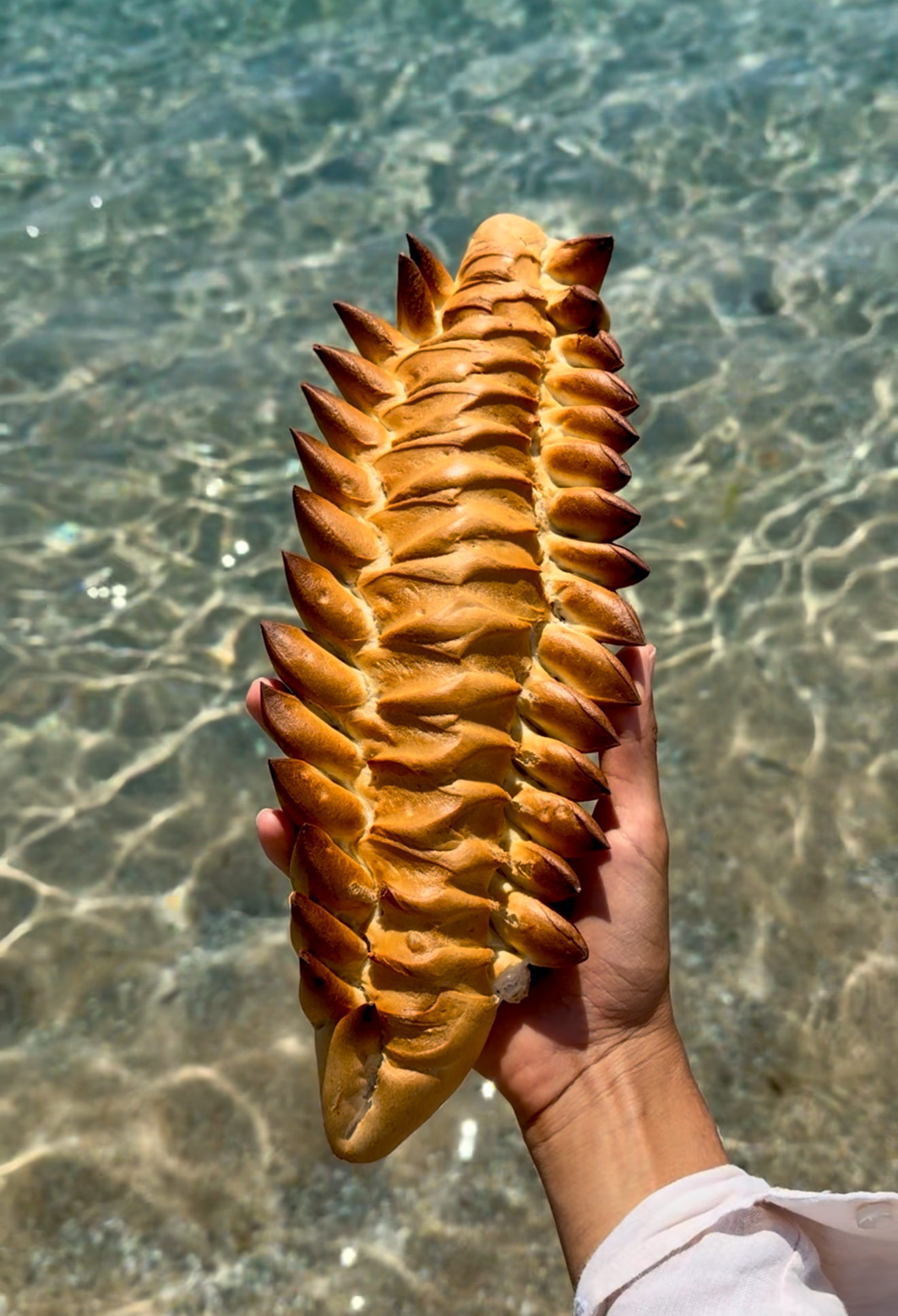 A hand displays a golden loaf with leaf patterns and sharp edges, hovering over clear sunlit water; cuts mimic fish scales or leaves.