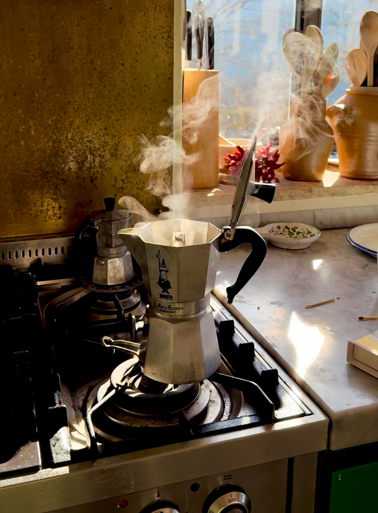Una moka prepara il caffè su un fornello a gas con il vapore, la luce del sole su utensili, piante in vaso e un piatto vicino alla finestra della cucina.