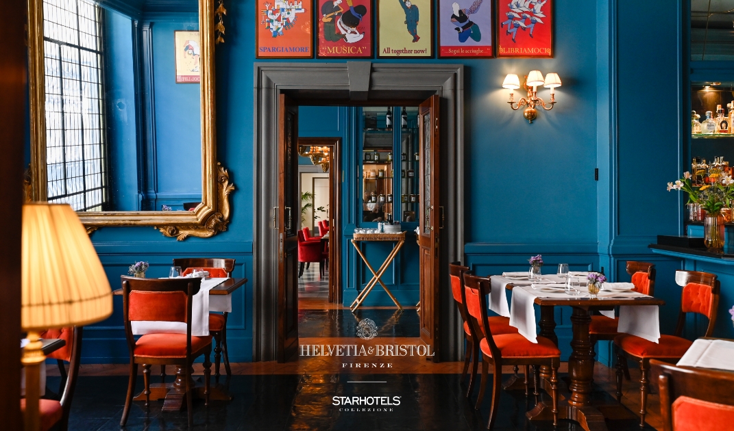 Elegant restaurant interior with blue walls, vintage mirrors, posters, white-tableclothed tables, and a bar visible through an open doorway.
