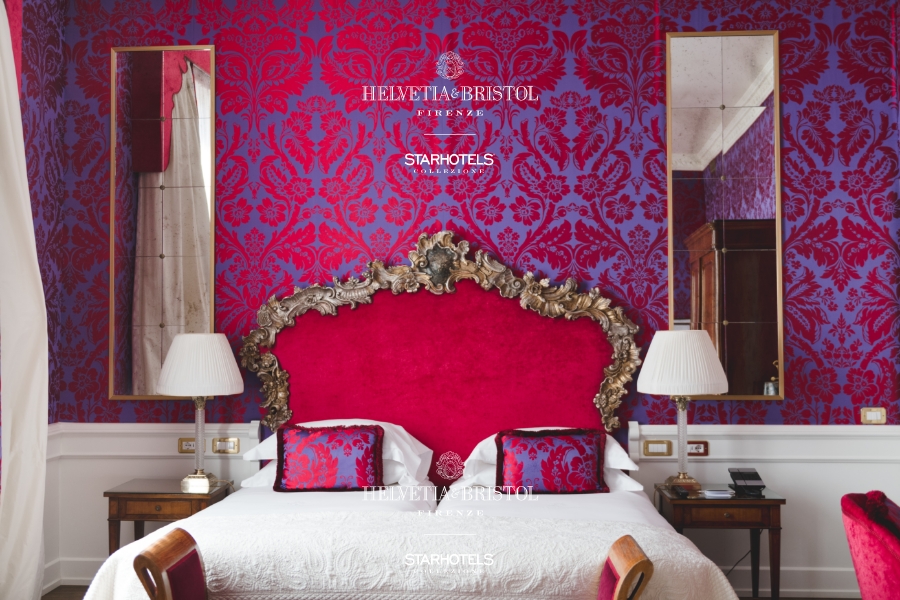 Luxurious hotel room - Ornate red velvet headboard, white bedding, floral wallpaper, mirrored bedside tables with lamps, classic décor.