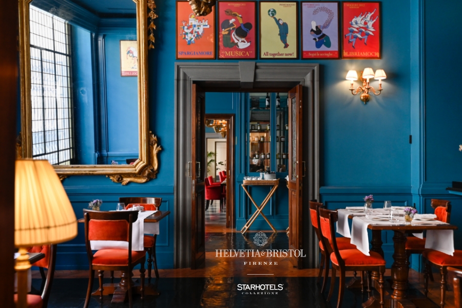 Elegant restaurant with blue walls, gold mirrors, red chairs, white tablecloths; posters and logos visible. Stylish adjoining room.
