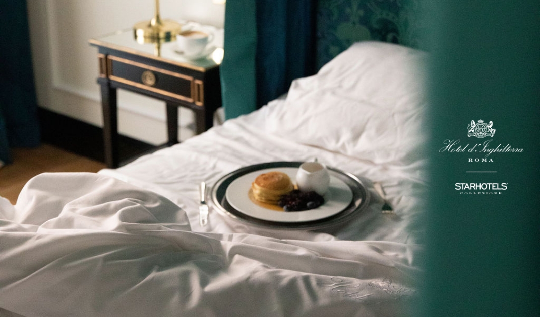 A breakfast tray with pancakes, syrup, jam, coffee on a rumpled white-sheeted hotel bed; visible hotel logos in soft light.