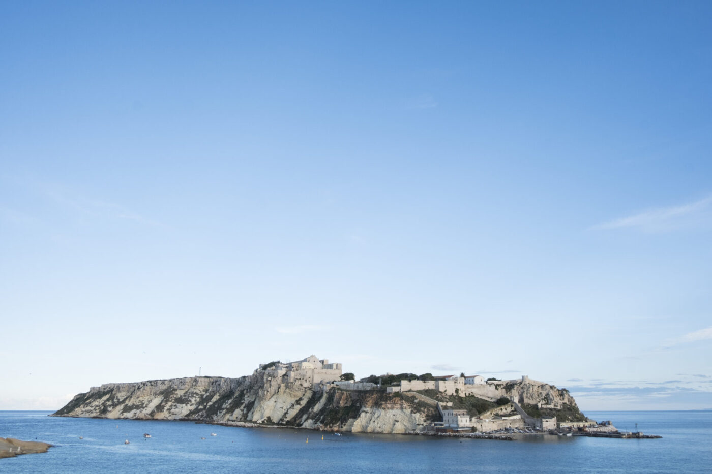 A rocky island dotted with ancient stone buildings and forts rises from tranquil blue water beneath a cloudless sky.