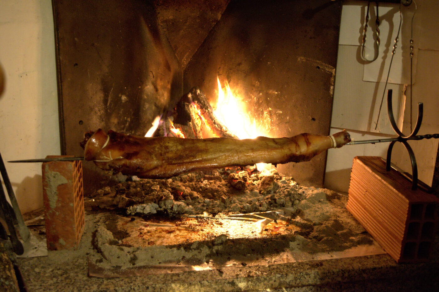 A whole roast lamb skewered and cooking over glowing embers and flames in a rustic fireplace.