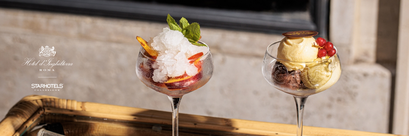Two stylish glass cups on a wooden tray outside: one with ice, peach, mint; the other with gelato, currants, chocolate coin in sunlight.