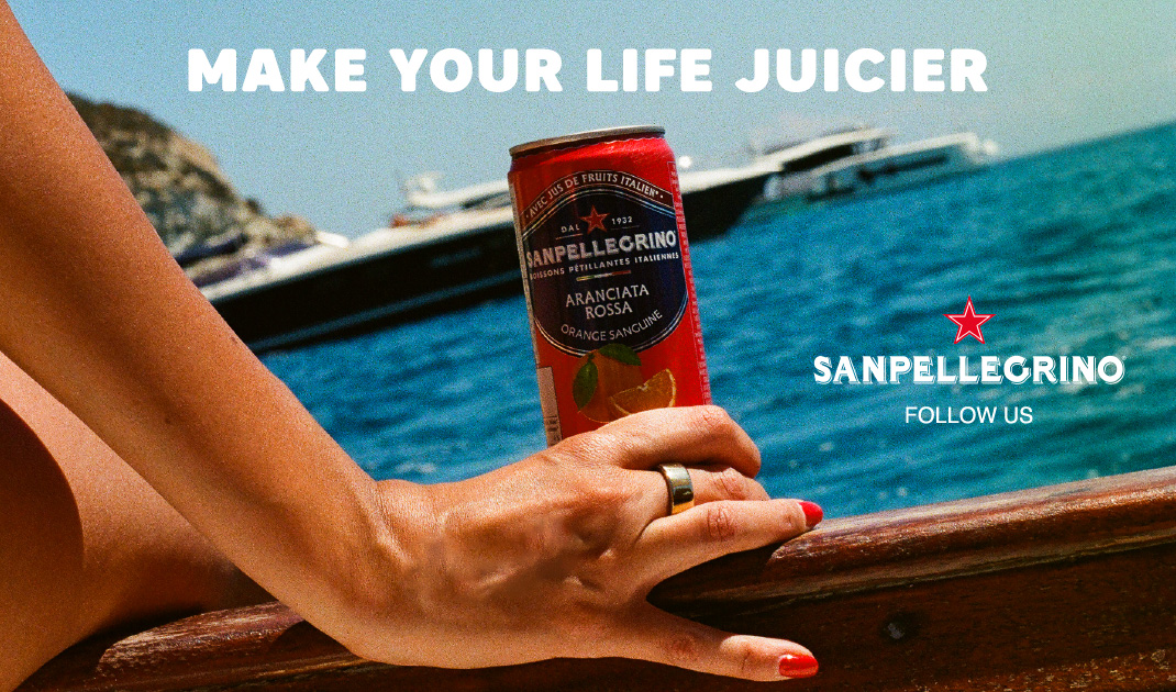 Una mano con le unghie rosse tiene una lattina di Sanpellegrino su una barca; sono visibili il mare, gli yacht e la scritta "MAKE YOUR LIFE JUICIER".
