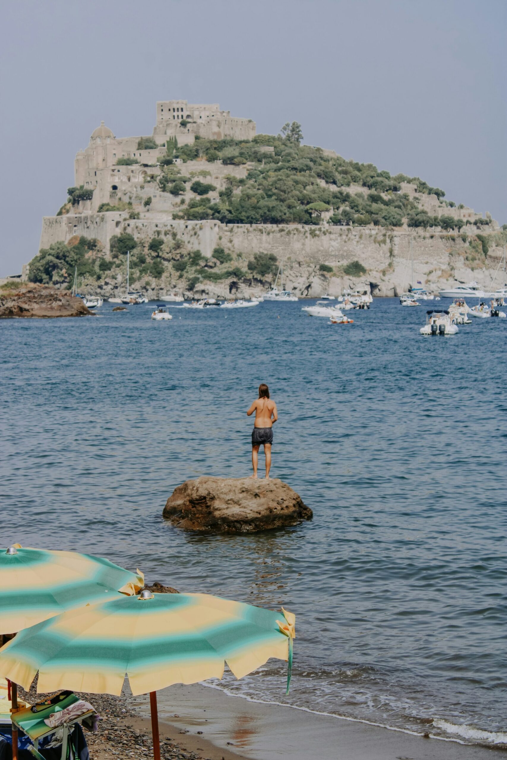[Una persona si trova su una roccia nel mare, di fronte a un castello storico su una collina lussureggiante. Ombrelloni e barche colorate sono visibili in primo piano e sullo sfondo, creando una scena costiera estiva.