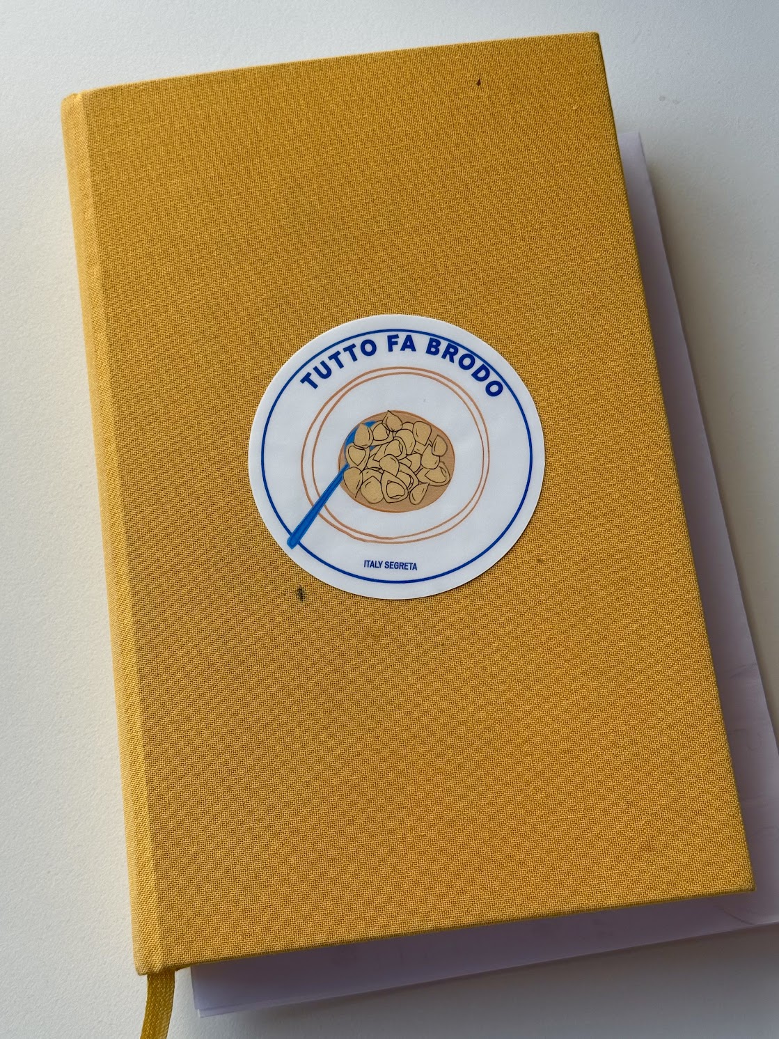 TUTTO FA BRODO Sticker on Yellow Notebook - A yellow hardcover notebook with a TUTTO FA BRODO bowl of pasta sticker from the A Tavola Sticker Pack on the cover, shown lying on a white surface with part of a sheet of paper visible underneath.