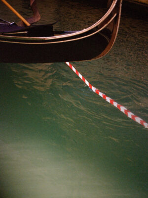 The front of a gondola floats on green water, facing a red and white striped barrier stretching across the canal.