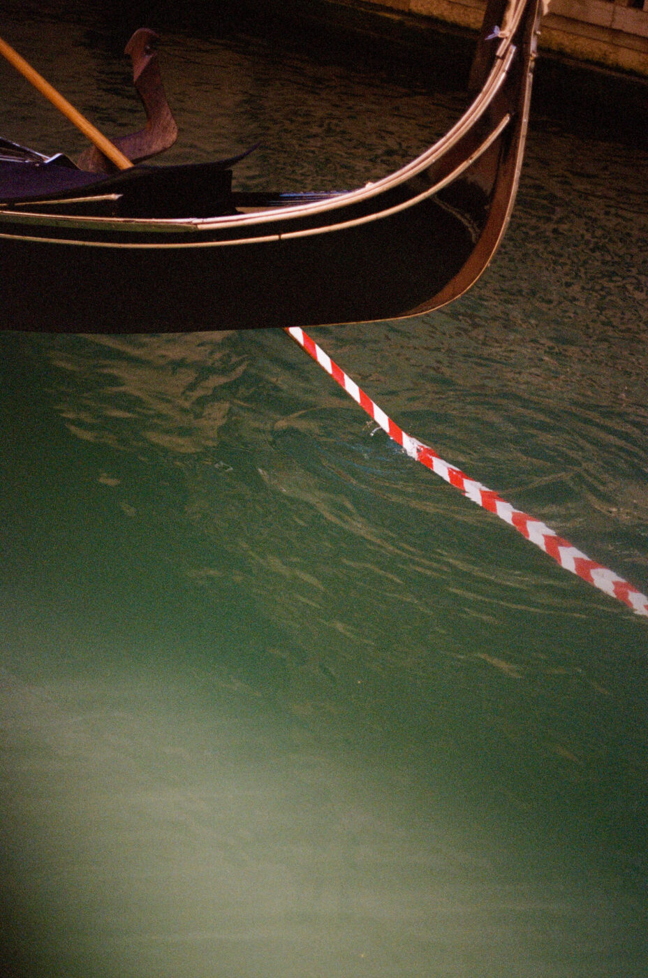 The front of a gondola floats on green water, facing a red and white striped barrier stretching across the canal.