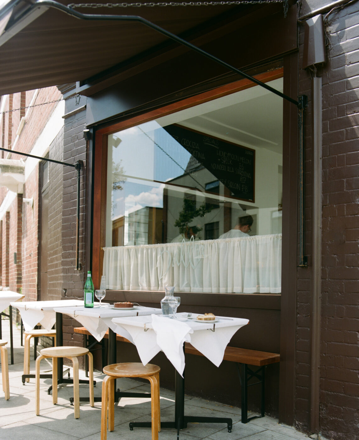 A sidewalk café with white-clothed tables, set with plates, glasses, green bottle; wooden stools and a window reflecting buildings.