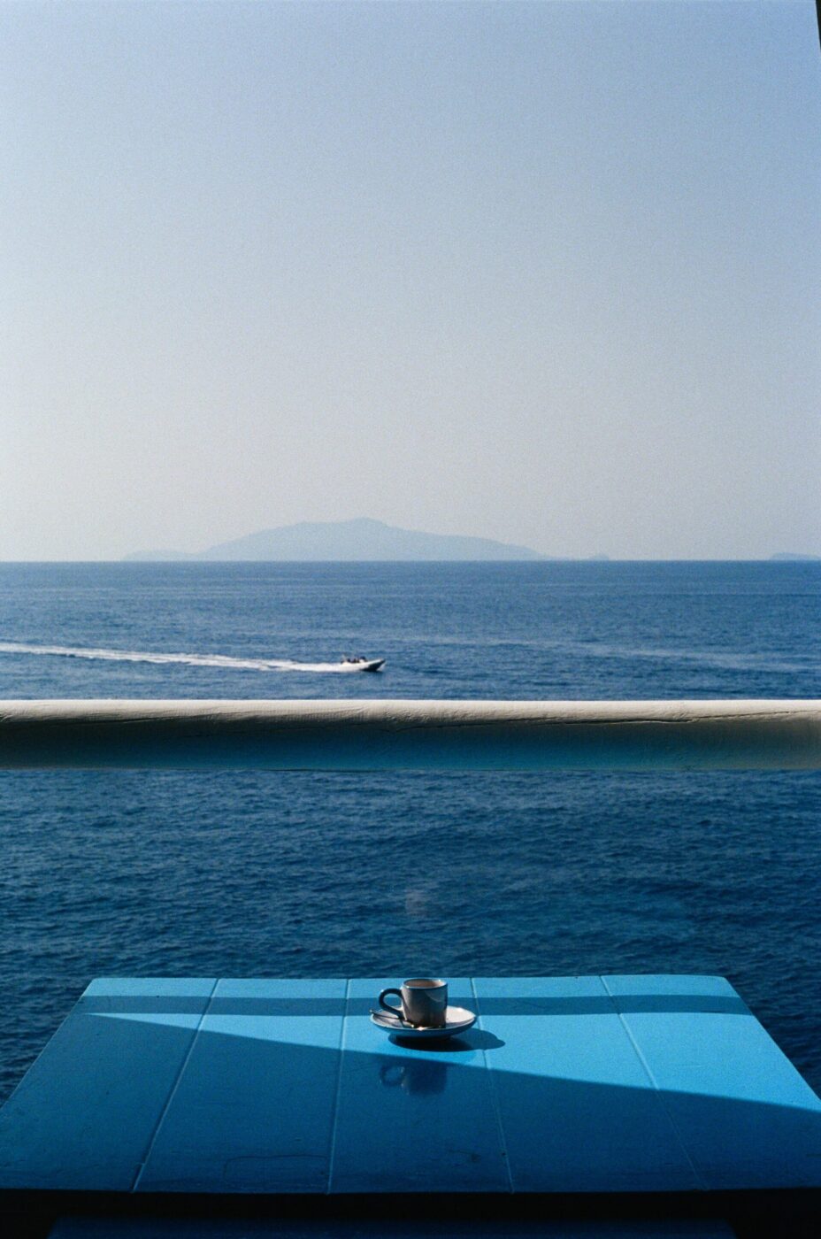 Una tazza di caffè su un tavolo azzurro che si affaccia su un mare calmo, con una barca che sfreccia e un'isola lontana sotto un cielo limpido.