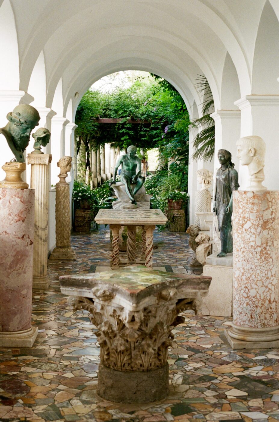Un corridoio di marmo con soffitti ad arco, busti classici, statue lungo i lati e pavimento a mosaico conduce a un giardino lussureggiante.