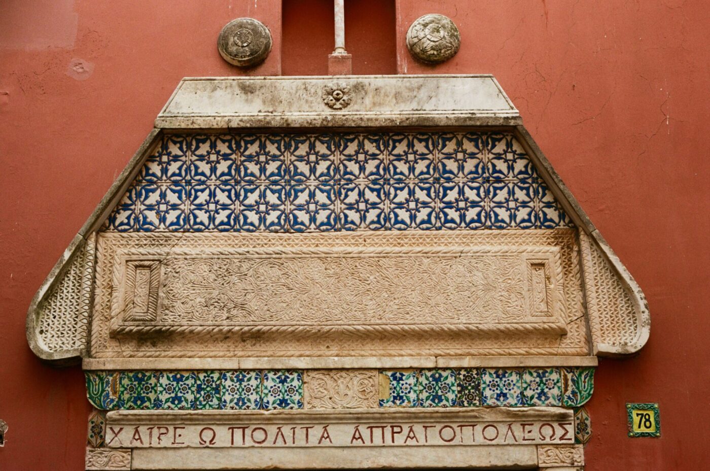 Targa in pietra ornata con testo greco, intagli, piastrelle bianche e blu su una parete rossa; due dettagli rotondi e 78 si trovano sopra/destra.