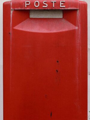 Primo piano di una cassetta postale rossa con l'etichetta POSTE in bianco, su uno sfondo semplice. La cassetta presenta segni di usura e di utilizzo.