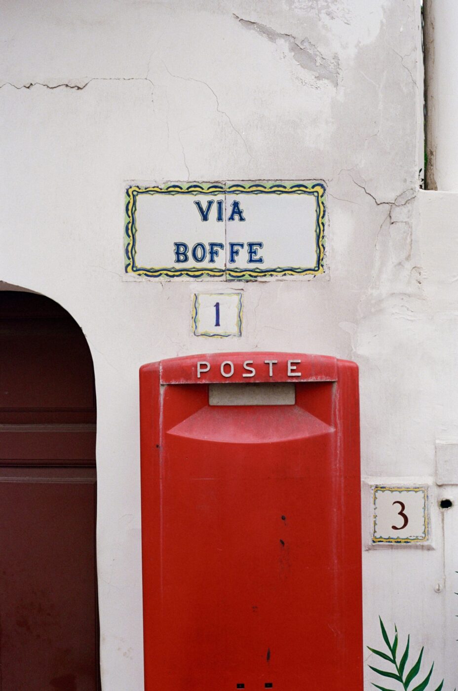 [Titolo immagine] - Una cassetta postale rossa POSTE su un muro bianco sotto l'insegna di Via Boffe 1, con una targa con il numero 3 e una pianta verde nelle vicinanze.