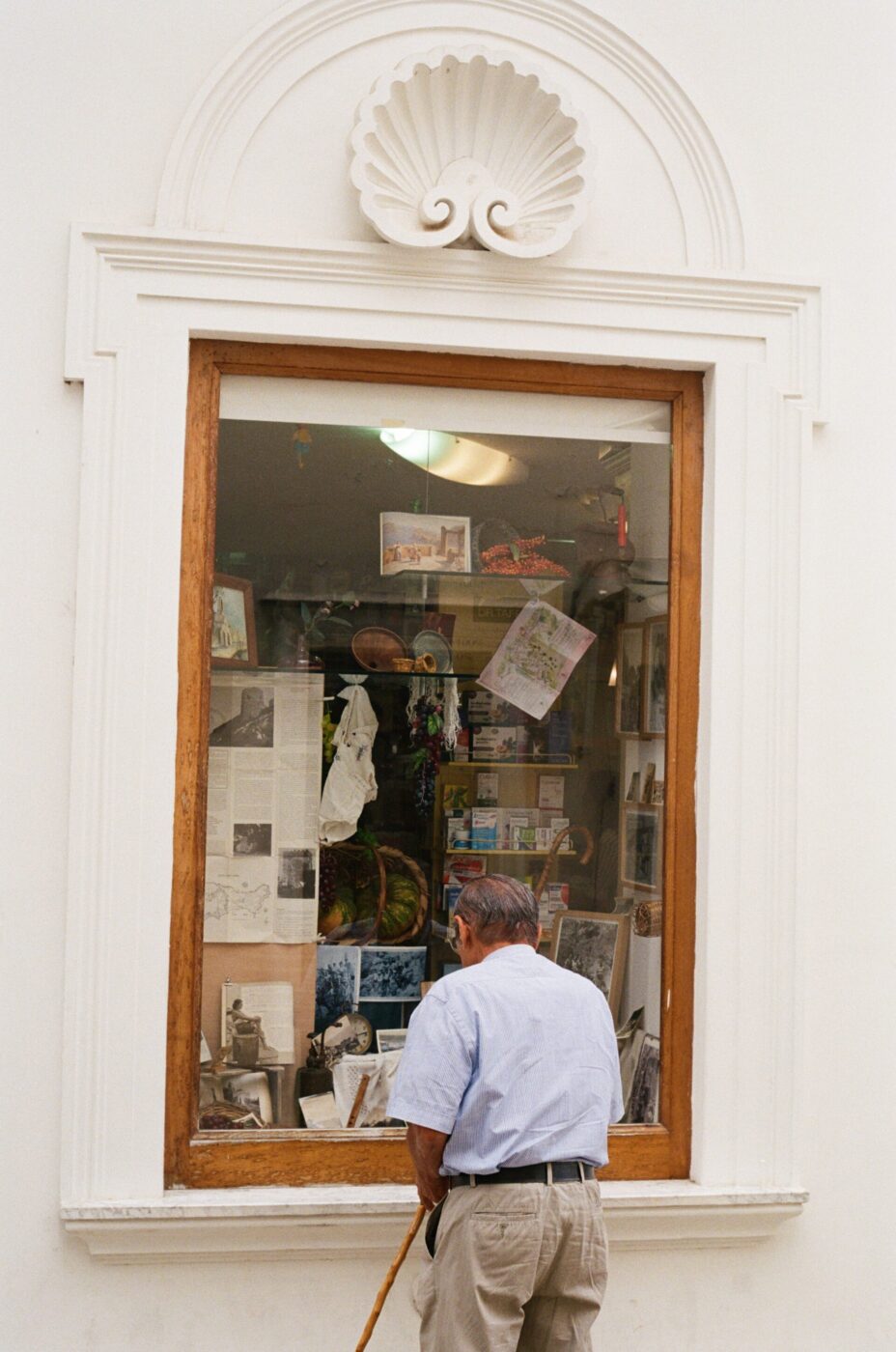 Un uomo anziano con un bastone si trova accanto a una vetrina di libri, cartoline e souvenir in una parete bianca con decorazioni di conchiglie sopra la cornice.