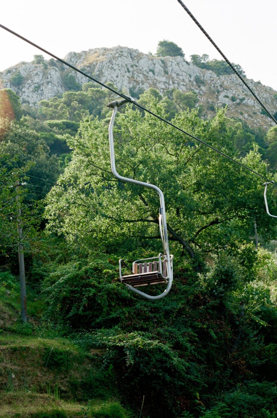 Una seggiovia vuota scivola sopra alberi e cespugli verdi, con un pendio roccioso e alberi sparsi sullo sfondo sotto un cielo limpido.