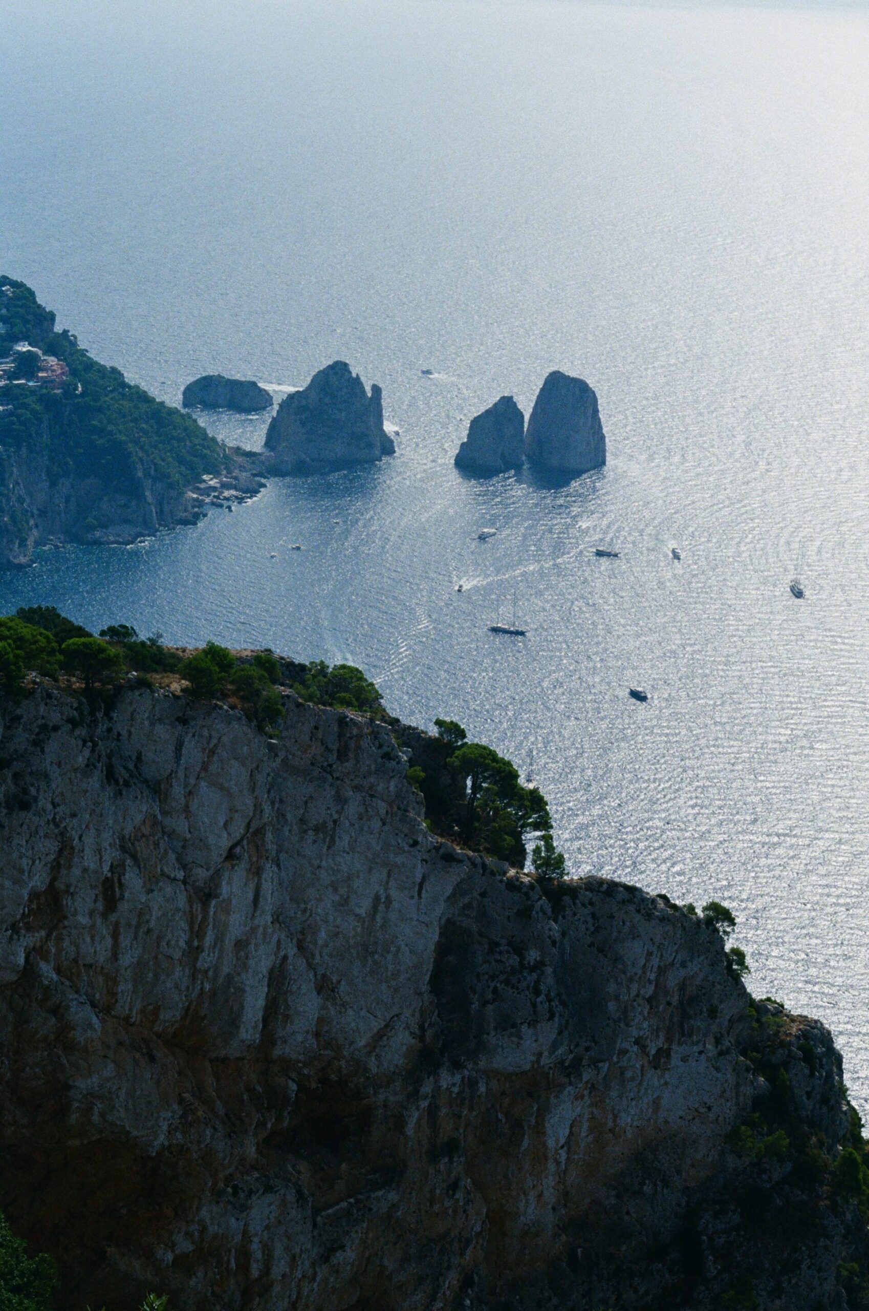 Vista di ripide scogliere rocciose con poca vegetazione sopra un mare azzurro e calmo; tre Faraglioni emergono dall'acqua, piccole barche nelle vicinanze.