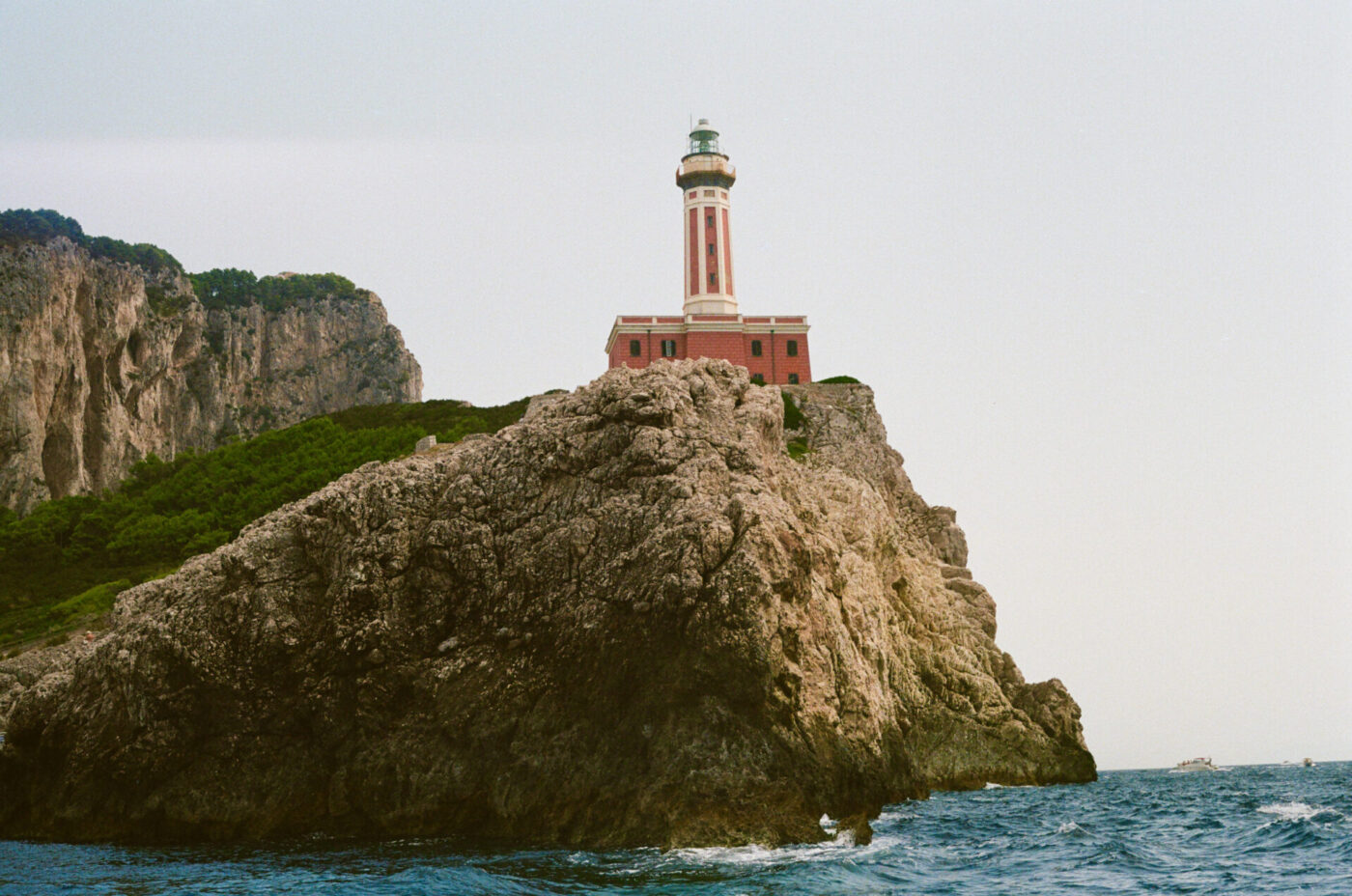 Un faro rosso e bianco si erge su una scogliera rocciosa in riva al mare, con colline verdi, cielo azzurro e onde che si infrangono sugli scogli sottostanti.