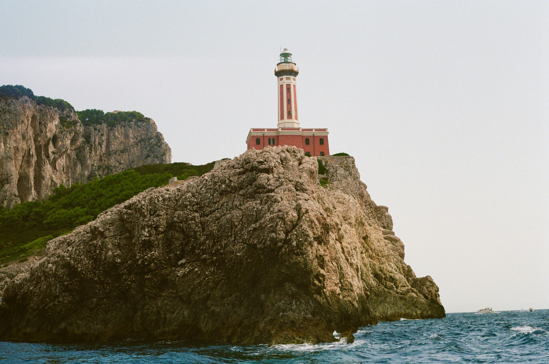 Un faro rosso e bianco si erge su una scogliera rocciosa in riva al mare, con colline verdi, cielo azzurro e onde che si infrangono sugli scogli sottostanti.