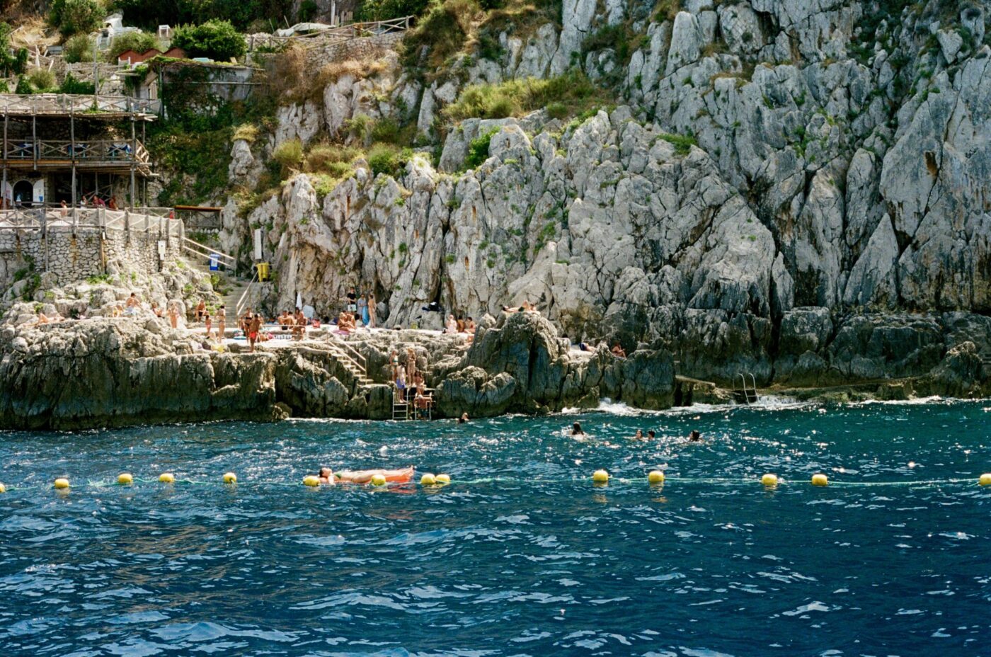 Le persone si rilassano sulle scogliere rocciose in riva al mare; i bagnanti galleggiano vicino a boe gialle, con edifici rustici sulla riva e fogliame verde nelle vicinanze.