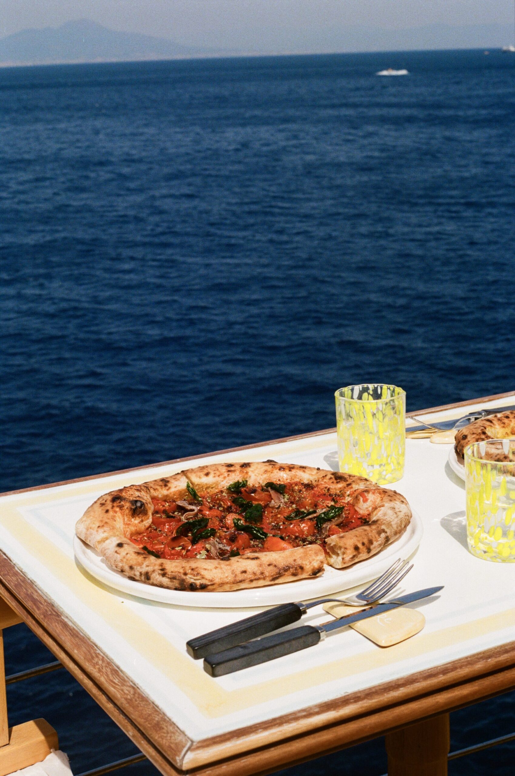 Una pizza cotta a legna e due bevande sono appoggiate su un tavolo con vista sul blu intenso del mare e sulle montagne lontane sotto un cielo limpido.