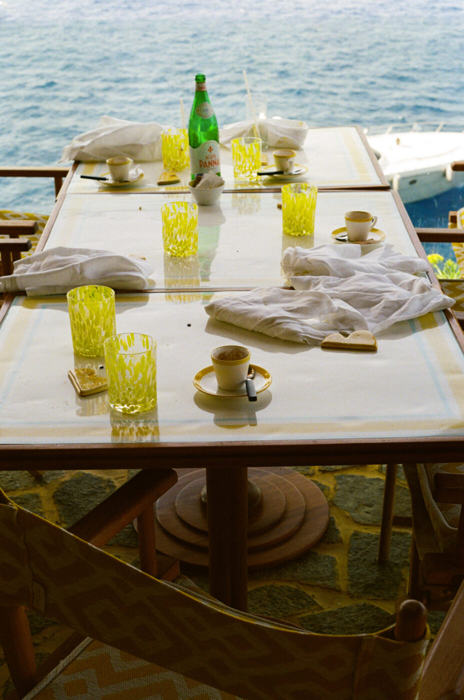 Tavolo all'aperto in riva al mare con bicchieri gialli vuoti, tazze da caffè, una bottiglia verde, tovaglioli e una barca sull'acqua sul retro.