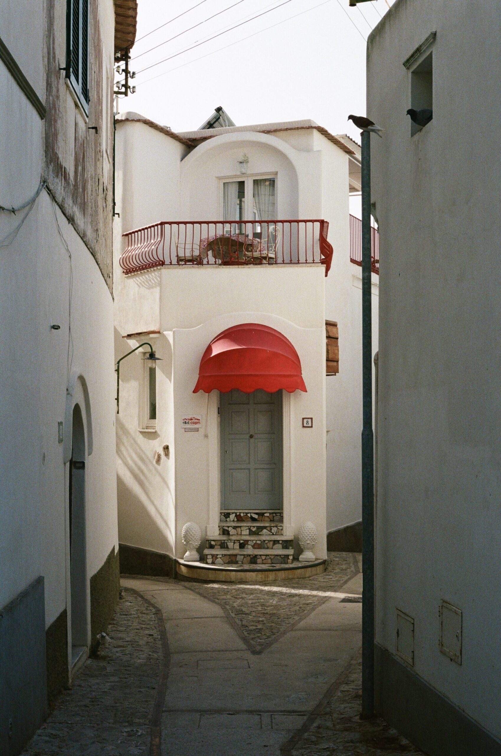 Uno stretto vicolo conduce a un edificio bianco con finestre ad arco, tenda rossa e balcone; la luce del sole crea ombre delicate.